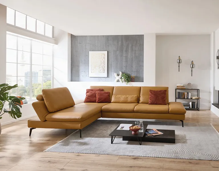 Großes Ecksofa in Cognacfarbe mit roten Kissen in einem hellen Wohnzimmer, dekoriert mit Teppich, Couchtisch, Wandbild und großen Fenstern für viel Tageslicht.